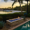 Tropical poolside setting with a rectangular stone fire pit filled with blue fire glass, glowing beautifully against the backdrop of palm trees and a tranquil waterfront sunset.