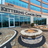 Commercial-grade fire pit with glass wind guard, powered by HPC Penta burner, glowing with vibrant flames outside the Kettering Cancer Center—creating a welcoming outdoor ambiance.