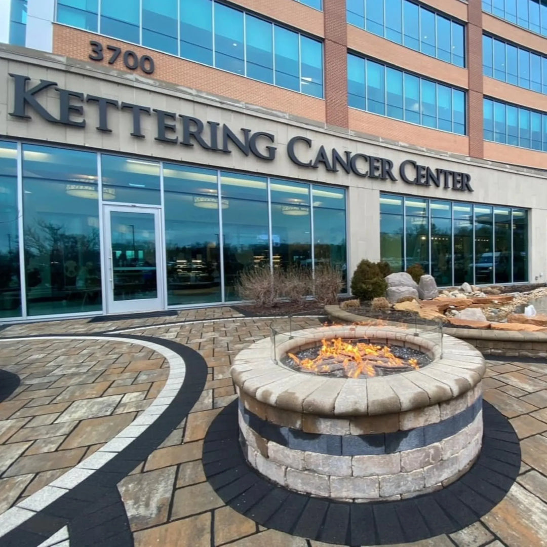 Commercial-grade fire pit with glass wind guard, powered by HPC Penta burner, glowing with vibrant flames outside the Kettering Cancer Center—creating a welcoming outdoor ambiance.