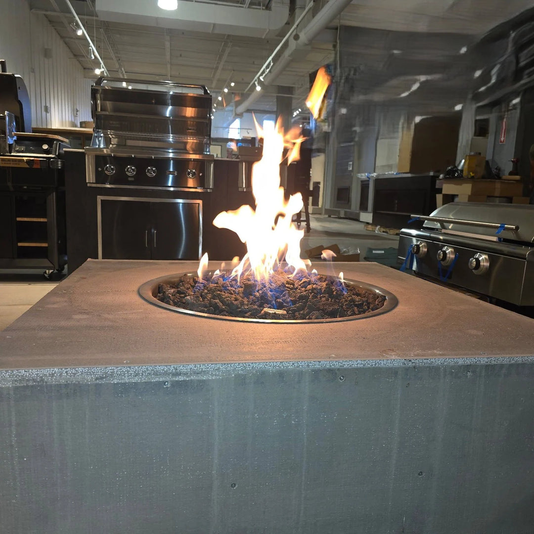 An HPC fire pit setup in a showroom, with a square concrete top and dark lava rock showcasing bold yellow-orange flames – perfect for modern outdoor kitchen designs.