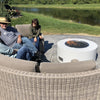 A white modern round fire pit with black fire glass and an HPC Penta burner glows gently as a group relaxes outdoors by a scenic pond, creating a cozy atmosphere.