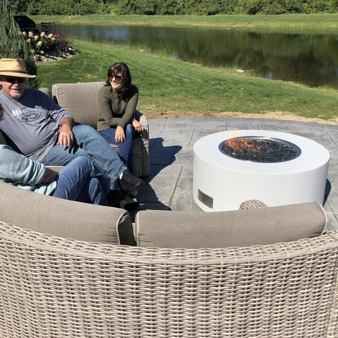A white modern round fire pit with black fire glass and an HPC Penta burner glows gently as a group relaxes outdoors by a scenic pond, creating a cozy atmosphere.