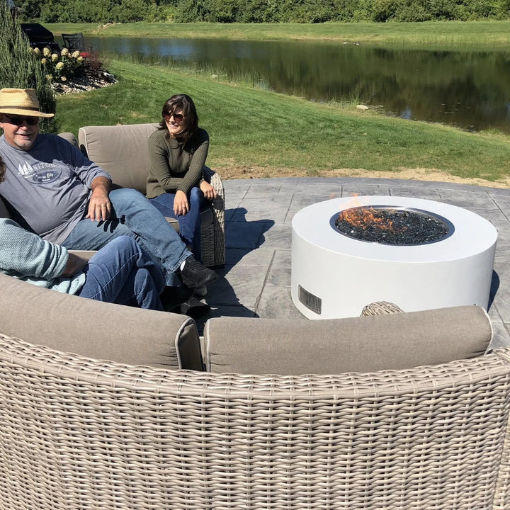 A white modern round fire pit with black fire glass and an HPC Penta burner glows gently as a group relaxes outdoors by a scenic pond, creating a cozy atmosphere.