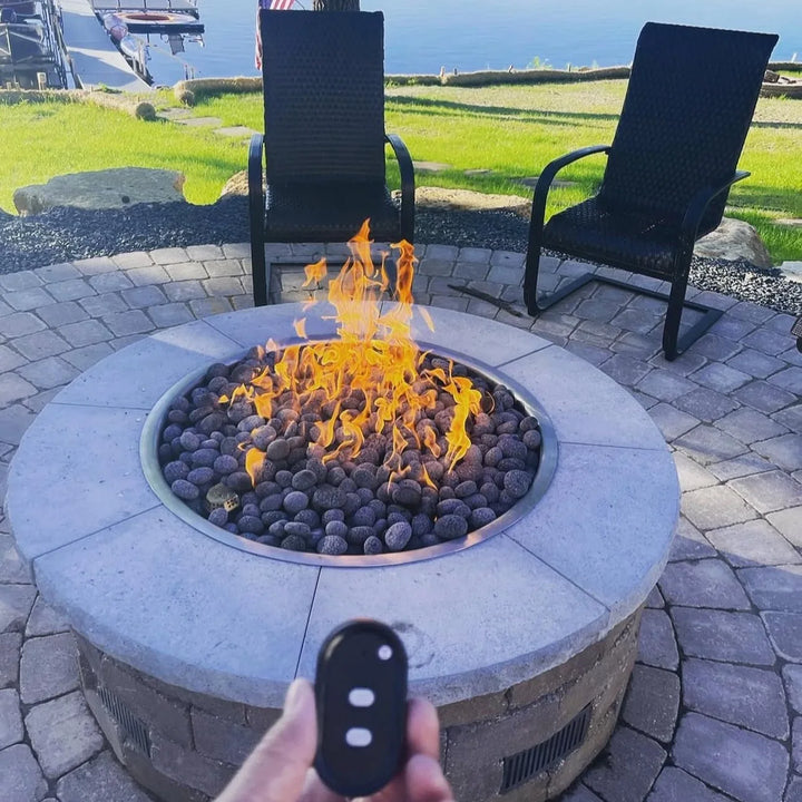 Close-up of a hand holding an HPC remote in front of a round paver fire pit filled with lava rock, flames dancing high thanks to the powerful HPC Penta burner.