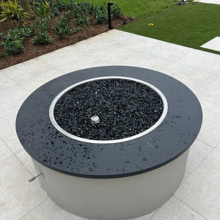 Sleek round fire pit with black granite top and wet reflective black fire glass, displaying HPC’s stainless steel drop-in burner ring in an elegant outdoor setting.