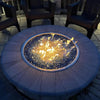 A glowing round fire pit filled with shimmering blue fire glass on a stone paver base, surrounded by chairs for cozy evening relaxation under the stars.