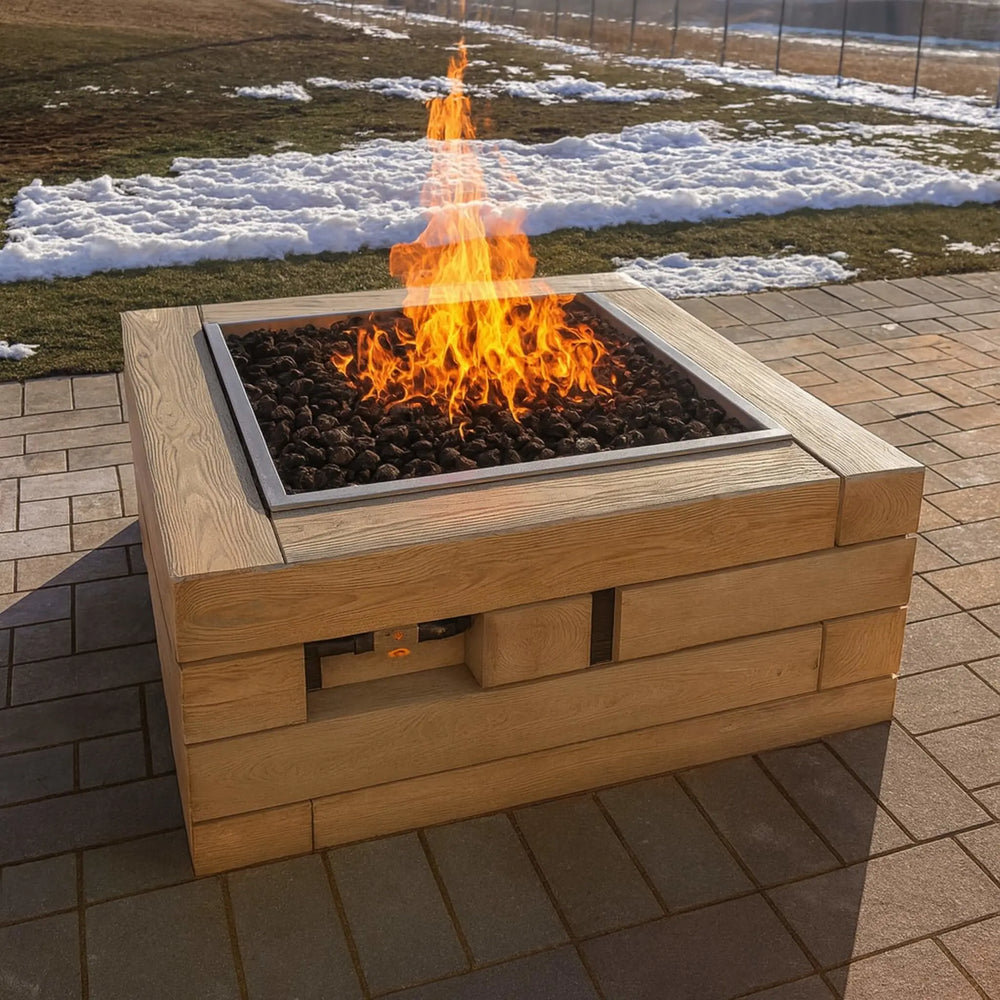 A wood-look outdoor fire pit with a Square Drop-In Penta Burner by HPC Fire glows warmly with natural lava rock and tall dancing flames, set against a snowy lawn backdrop.