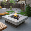 Modern backyard patio scene featuring a sleek square concrete fire pit with a vibrant flame rising from lava rock media. Two glasses of red wine and a bottle rest on the wide ledge, creating an inviting and intimate ambiance / perfect for evening relaxation or romantic outdoor gatherings.