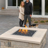 A smiling couple with their dog stands beside a custom-built patio fire pit featuring an HPC Fire Square Drop-In Pan with a Penta burner and black fire media, creating a warm and inviting backyard atmosphere.