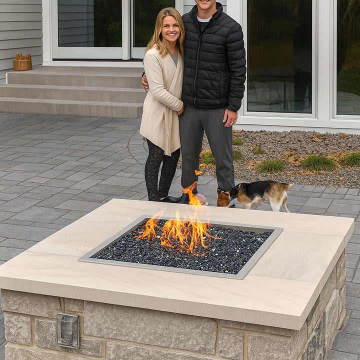 A smiling couple with their dog stands beside a custom-built patio fire pit featuring an HPC Fire Square Drop-In Pan with a Penta burner and black fire media, creating a warm and inviting backyard atmosphere.