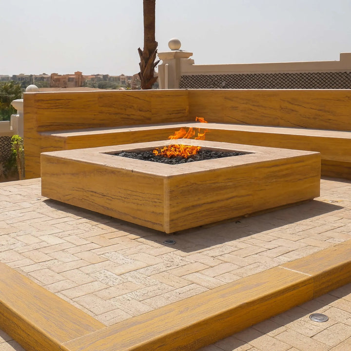 Modern sun-drenched outdoor seating area featuring a recessed square gas fire pit built into a stone platform, powered by an HPC square flat burner, radiating warmth with bright flames and black lava rocks.