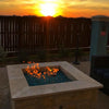Square stone gas fire pit filled with shimmering blue fire glass, flames dancing warmly against a glowing sunset in a cozy backyard setting.