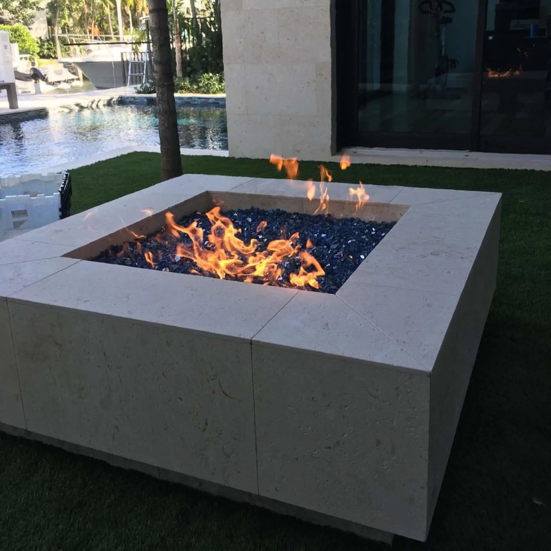 Modern square fire pit with beige stone finish and sparkling blue fire glass, set beside a contemporary pool for a sleek outdoor retreat.