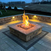Square stone fire pit ignited with bright, vertical flames at dusk, surrounded by built-in stone seating and soft landscape lighting / an ideal backyard retreat powered by HPC's square flat burner system.