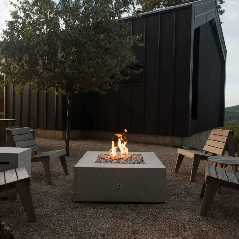 Scenic outdoor lounge with rustic wooden chairs encircling a concrete square fire pit filled with decorative pebbles / flames dancing atop an HPC Fire square flat burner for an inviting wilderness escape.