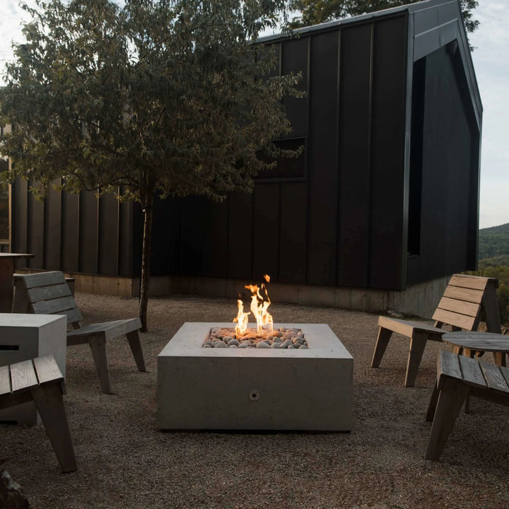 Scenic outdoor lounge with rustic wooden chairs encircling a concrete square fire pit filled with decorative pebbles / flames dancing atop an HPC Fire square flat burner for an inviting wilderness escape.