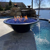 Sleek black GFRC Cazo fire and water bowl by The Outdoor Plus shown installed on a luxury poolside patio, with vibrant blue fire glass and flames dancing above. A calming water feature flows from the bowl into a sparkling swimming pool, creating a stunning visual contrast. Additional matching fire bowls glow in the background near a natural stone waterfall, all set against a peaceful lakeside view. Ideal for high-end outdoor living spaces combining fire, water, and modern design.