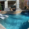 A stunning backyard scene featuring The Outdoor Plus Cazo Fire and Water Bowl in sleek Black GFRC, installed above a natural stone wall with a soothing water spill into a crystal-clear swimming pool. Bright orange flames dance in the bowl, creating a captivating contrast against the cool blue water. Sleek in-pool lounge chairs invite relaxation, while a cozy patio with wicker seating and a festive Christmas tree adds seasonal warmth to this modern outdoor retreat.