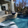 A luxurious backyard oasis featuring The Outdoor Plus Cazo Fire and Water Bowl in Black GFRC, elegantly perched on a stone-clad ledge. The fire bowl displays glowing fire glass and a graceful water flow into the sparkling blue pool below. In-pool loungers and a cozy covered patio with woven lounge chairs create the perfect space for relaxation and entertaining. Surrounded by natural stone, lush trees, and cascading waterfalls, this setup blends fire, water, and comfort in a modern outdoor retreat.