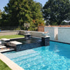 A beautifully designed modern backyard pool showcasing The Outdoor Plus Cazo Fire and Water Bowls in Rustic White GFRC, installed atop sleek columns. Each bowl features a glowing fire and a tranquil water spill that flows into the pool below. Surrounded by crisp blue water, dark mosaic tile spa walls, and lush green turf, this outdoor space offers a perfect mix of relaxation and elegance with a touch of resort-style ambiance.