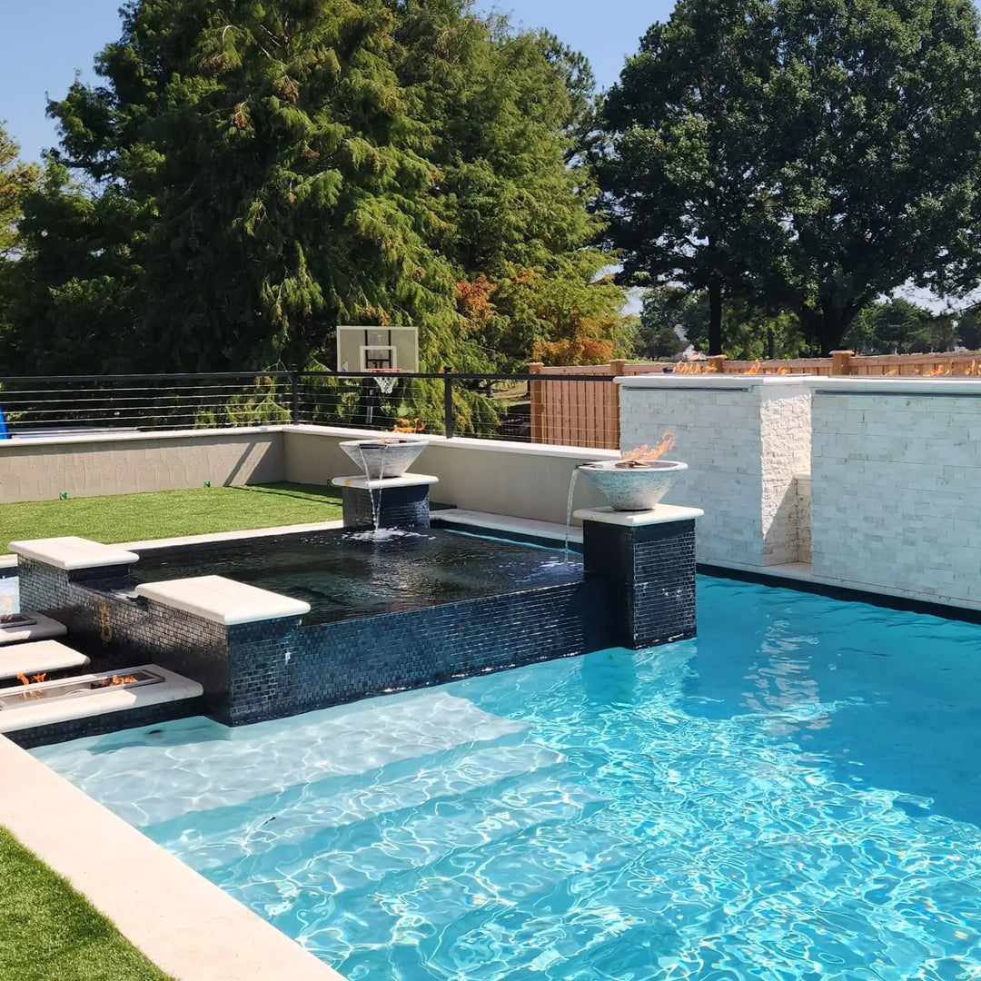 A beautifully designed modern backyard pool showcasing The Outdoor Plus Cazo Fire and Water Bowls in Rustic White GFRC, installed atop sleek columns. Each bowl features a glowing fire and a tranquil water spill that flows into the pool below. Surrounded by crisp blue water, dark mosaic tile spa walls, and lush green turf, this outdoor space offers a perfect mix of relaxation and elegance with a touch of resort-style ambiance.