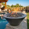 The Outdoor Plus Cazo Fire Bowl in a stunning Silver Vein powder-coated aluminum finish, beautifully displayed poolside on a raised pedestal. Filled with dark lava rock spheres and centered around a stainless steel burner, this handcrafted fire bowl adds modern elegance and radiant warmth to a luxurious backyard retreat.