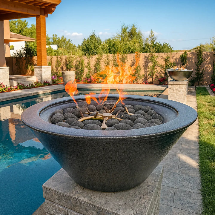 The Outdoor Plus Cazo Fire Bowl in a stunning Silver Vein powder-coated aluminum finish, beautifully displayed poolside on a raised pedestal. Filled with dark lava rock spheres and centered around a stainless steel burner, this handcrafted fire bowl adds modern elegance and radiant warmth to a luxurious backyard retreat.