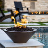 A cozy backyard scene showcasing The Outdoor Plus Cazo Round Fire Bowl in a smooth Chocolate GFRC finish. Set on a stone ledge beside a pool, the bowl is filled with natural black lava rock and features vibrant flames dancing in the evening light. In the background, dark Adirondack chairs and a matching fire feature add warmth and ambiance to this inviting outdoor space.