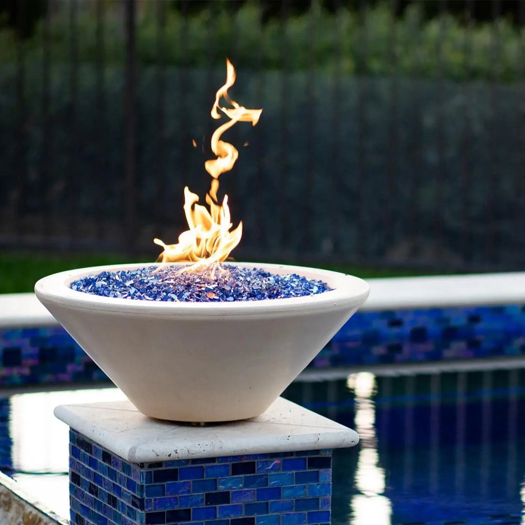 A clean and elegant poolside setup featuring The Outdoor Plus Cazo Round Fire Bowl in a soft Vanilla GFRC finish. Perched on a tiled pedestal, the bowl is filled with brilliant blue fire glass and showcases a bright, twisting flame above a deep blue pool. The setting exudes a modern, tropical vibe perfect for upscale outdoor living.