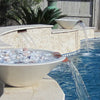 Contemporary limestone Cazo water bowls with decorative stones inside, spilling clear streams of water into a swimming pool for a luxury outdoor design.
