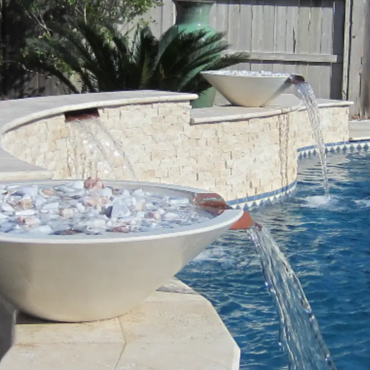 Contemporary limestone Cazo water bowls with decorative stones inside, spilling clear streams of water into a swimming pool for a luxury outdoor design.