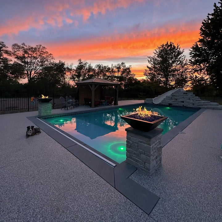A vibrant backyard at sunset showcasing The Outdoor Plus Maya Fire Bowl in black, glowing with fire glass and flames atop a stacked stone pedestal. Reflected pool lighting and a nearby lounging dog complete this warm, inviting evening atmosphere.