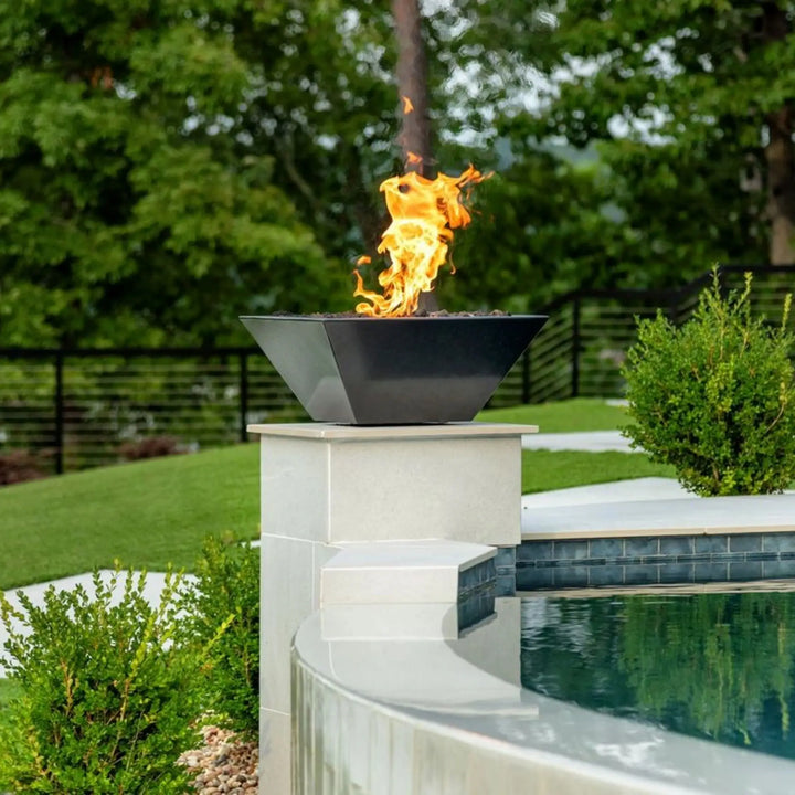 A sleek Maya Fire Bowl by The Outdoor Plus in black, proudly featured poolside atop a light stone pedestal. The tall, elegant flames create a striking contrast against the lush green landscape, blending modern design with outdoor serenity.