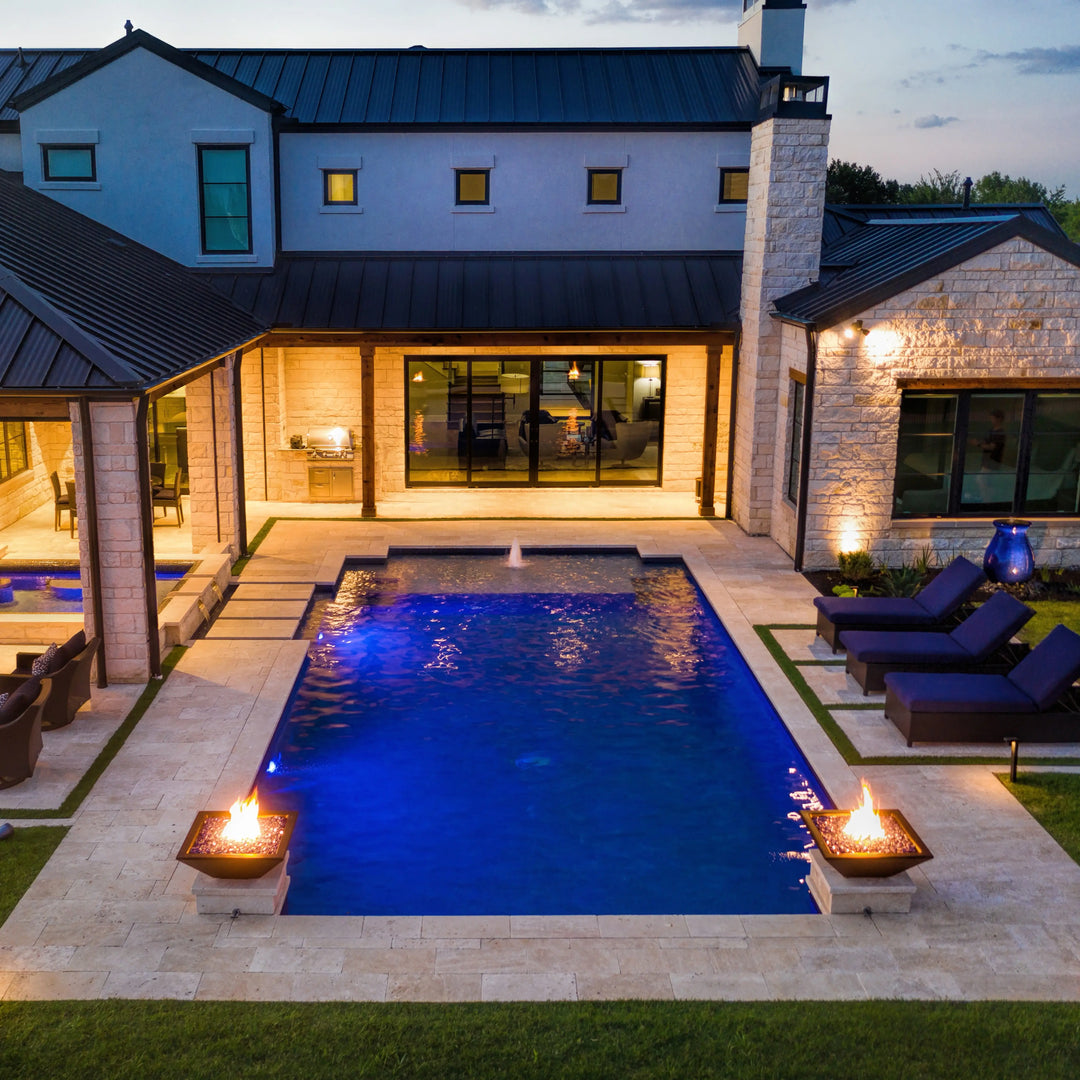 An elegant backyard view with two Maya Fire Bowls in black by The Outdoor Plus, perfectly flanking a modern pool. The glowing flames and ambient lighting create a welcoming atmosphere ideal for luxurious outdoor living.