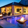 A cozy evening scene featuring a close-up of the Maya Fire Bowl in black finish from The Outdoor Plus, glowing with warm flames and fire glass. The bowl is set on a light stone pedestal beside a pool, surrounded by a softly lit patio lounge.