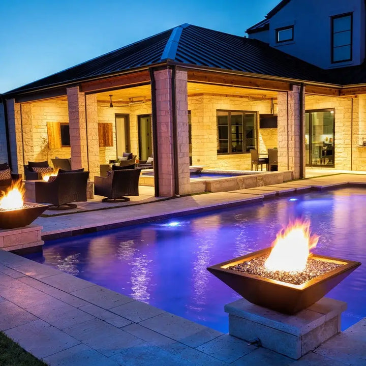 A cozy evening scene featuring a close-up of the Maya Fire Bowl in black finish from The Outdoor Plus, glowing with warm flames and fire glass. The bowl is set on a light stone pedestal beside a pool, surrounded by a softly lit patio lounge.