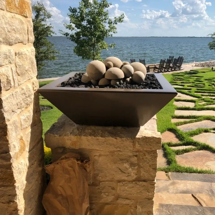 A Chocolate-finished Maya Fire Bowl by The Outdoor Plus is beautifully staged on a stone pillar with a lakefront view. Filled with smooth ceramic fire stones and black lava rock, this elegant bowl adds sculptural charm and warmth to a serene outdoor patio space by the water.
