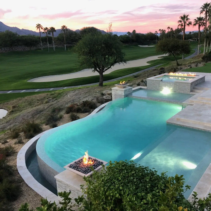 A breathtaking infinity-edge pool overlooking a golf course, featuring Maya Fire Bowls in Vanilla finish by The Outdoor Plus. The warm flames reflect against the water as palm trees and mountains complete the tranquil backdrop.