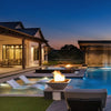A picturesque backyard view at dusk with Maya Fire Bowls in Vanilla finish from The Outdoor Plus, installed along the edge of a glowing modern pool. The flickering fire elements add elegance and warmth to this beautifully lit luxury setting.