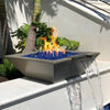 A stunning close-up of The Outdoor Plus Maya Fire and Water Bowl in a smooth Chestnut finish, set atop a white stone ledge near tropical foliage. Vibrant flames rise from blue fire glass as a soothing stream of water pours gracefully from the built-in scupper, offering a bold blend of heat and tranquility in this modern outdoor design.