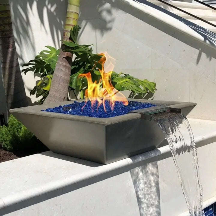 A stunning close-up of The Outdoor Plus Maya Fire and Water Bowl in a smooth Chestnut finish, set atop a white stone ledge near tropical foliage. Vibrant flames rise from blue fire glass as a soothing stream of water pours gracefully from the built-in scupper, offering a bold blend of heat and tranquility in this modern outdoor design.