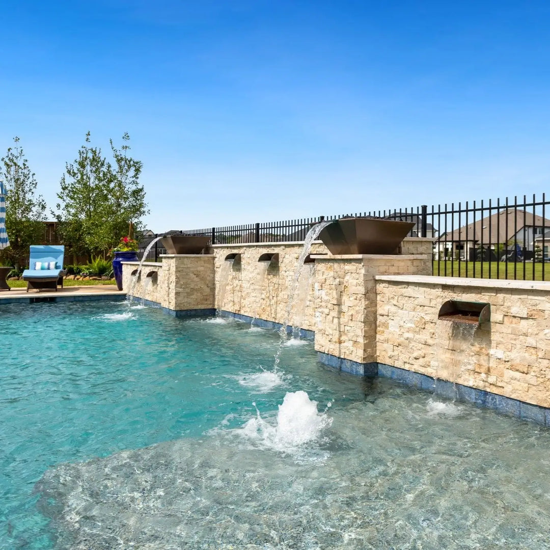 A luxurious backyard pool featuring multiple Maya Fire and Water Bowls by The Outdoor Plus in a modern Chocolate finish. Mounted on a beautifully crafted natural stone wall, each bowl spills a steady stream of water into the sparkling blue pool below. Bubblers and lounge seating in the shallow tanning ledge create a serene and resort-style atmosphere perfect for outdoor relaxation and upscale design.