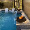 A luxurious poolside view featuring The Outdoor Plus Maya Fire and Water Bowls in a rich Chocolate finish. Set atop stylish mosaic pillars, the bowls feature glowing flames over black lava rocks with gentle water flows cascading into the blue pool. Sleek white in-pool loungers and lush landscaping create a serene, upscale backyard experience.