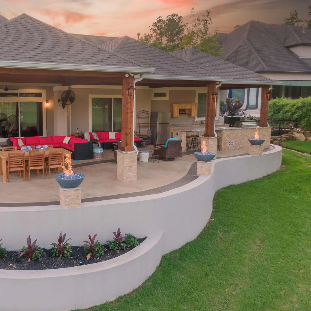 Gray Sedona Fire Bowls by The Outdoor Plus with blue fire glass, neatly positioned on stone columns across a lakefront patio, delivering breathtaking views and glowing warmth.