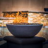 Modern rustic fire bowl with vivid flames dancing above black fire glass, set against a tranquil water feature backdrop. The Sedona Fire Bowl in a smooth rustic gray finish adds warmth and ambiance to any luxury outdoor space / perfect for poolside gatherings or evening relaxation.