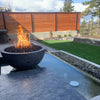 Rustic Gray Sedona Fire Bowl by The Outdoor Plus beautifully lit on a raised water feature, bringing warmth and elegance to a modern backyard patio space.
