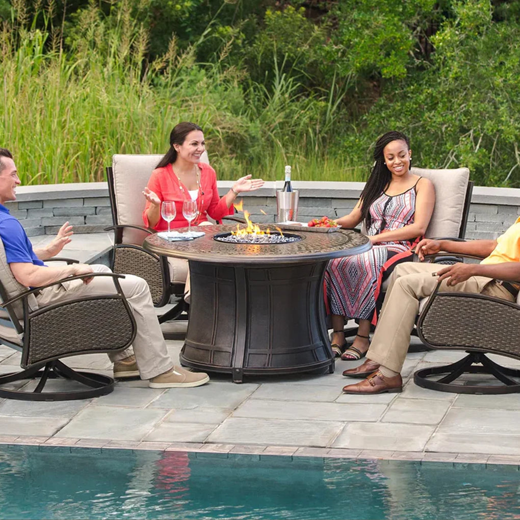 Group of four people seated around a round fire pit table on a stone patio next to a pool, engaged in conversation with drinks, surrounded by lush greenery