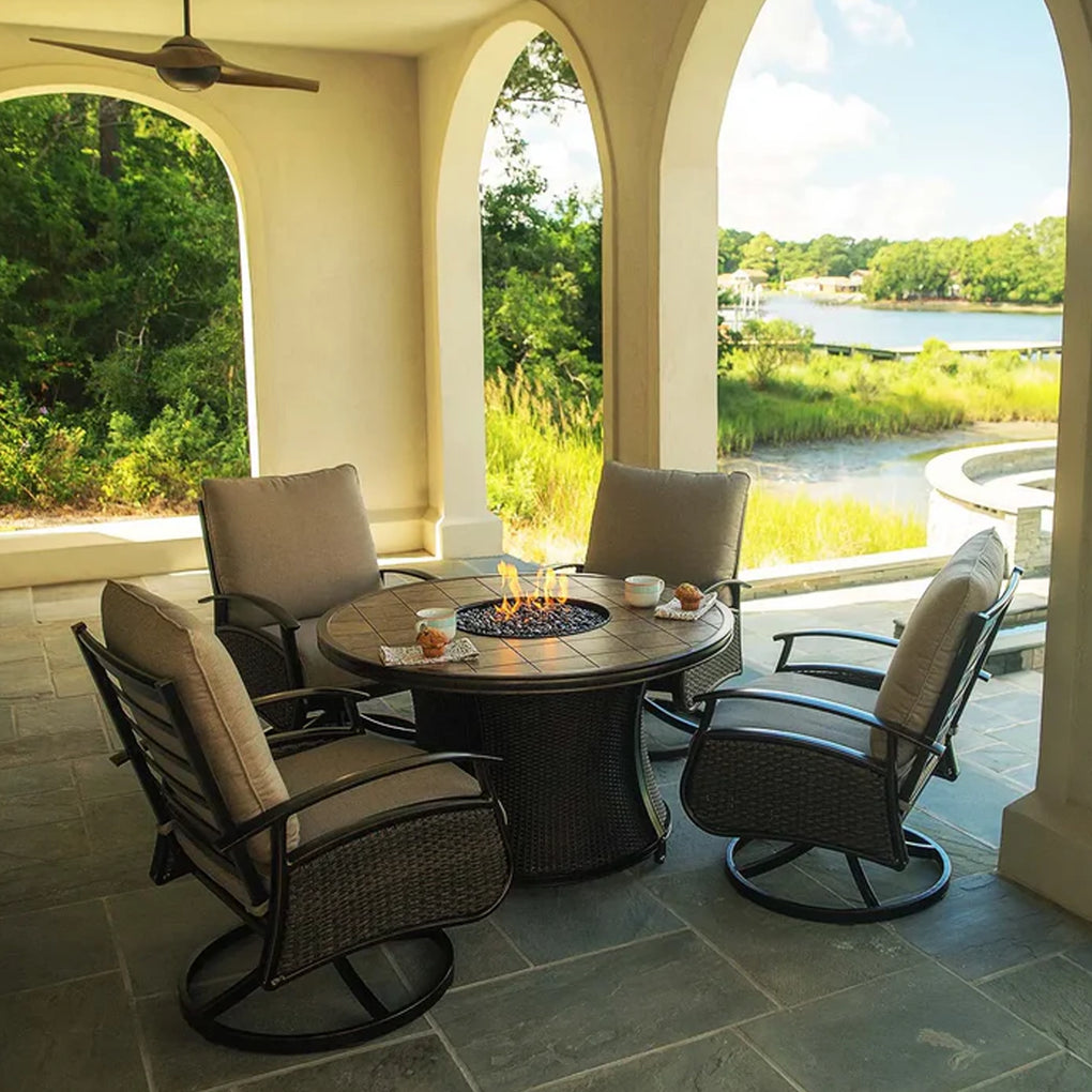 Outdoor patio seating area with a round fire pit table and four cushioned swivel chairs under a covered patio with archways, overlooking a scenic lake with trees