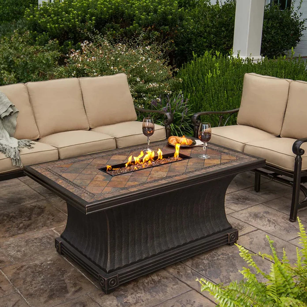 Outdoor patio with a rectangular tile-style fire pit table surrounded by two cushioned sofas, set against a backdrop of lush greenery, creating a cozy and inviting ambiance