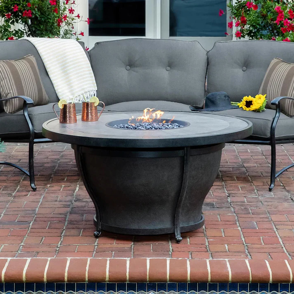 Outdoor seating area featuring a round fire pit table, tufted cushioned sofa, and armchairs on a brick patio, with decorative accessories and bright red flowers enhancing the cozy and inviting atmosphere